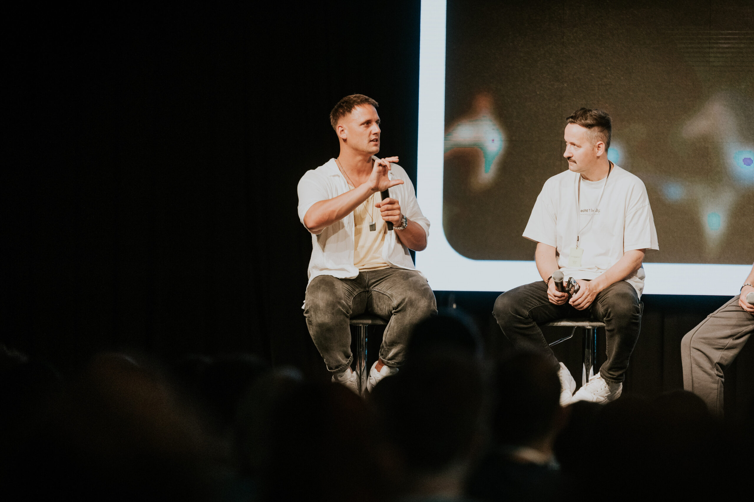Alt text: Two men are sitting on a stage, one is speaking and gesturing with his hand, while the other is listening.