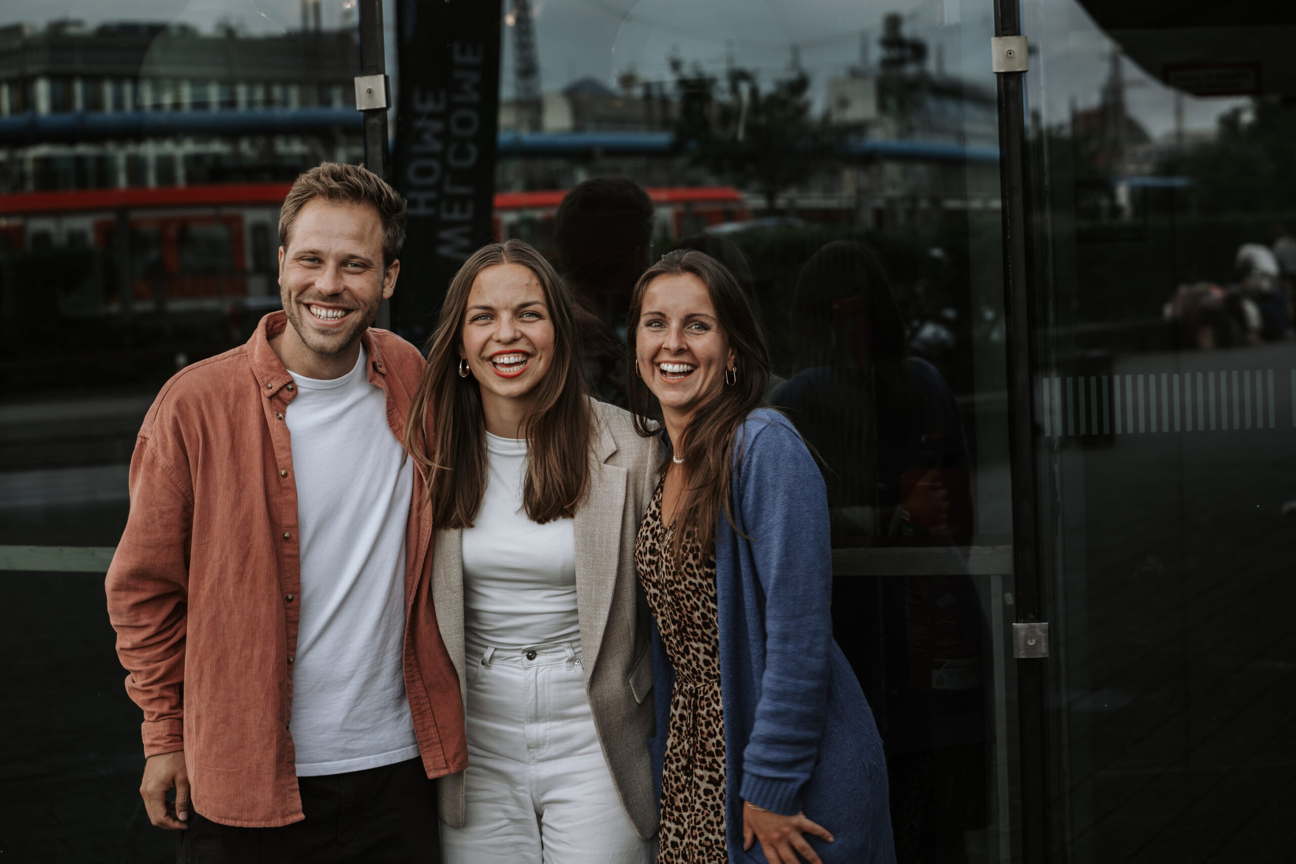 Drei Menschen stehen vor einem dunklen Glasgebäude, lachen und posieren für das Foto.