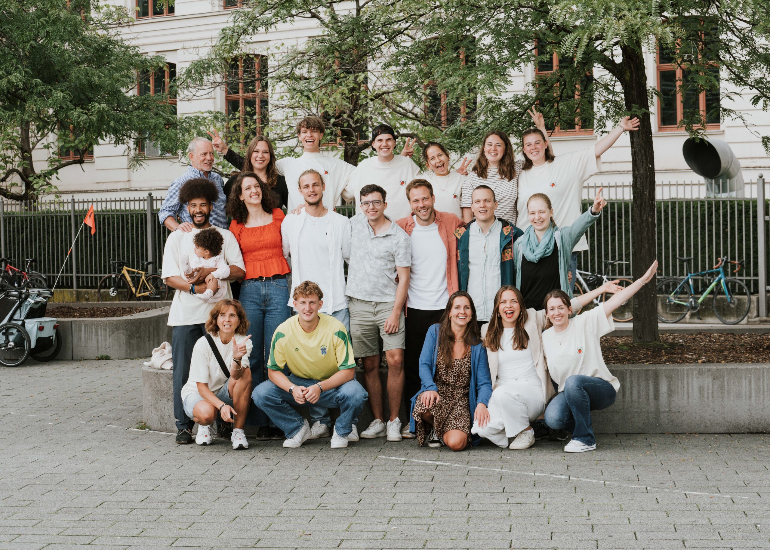 Mehrere Menschen stehen und sitzen im Freien vor einem Gebäude, einige winken.