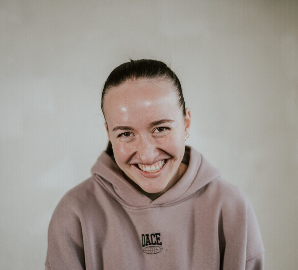 Junge Frau mit braunem Haar in einem hellbraunen Hoodie, lächelnd, vor neutralem Hintergrund.