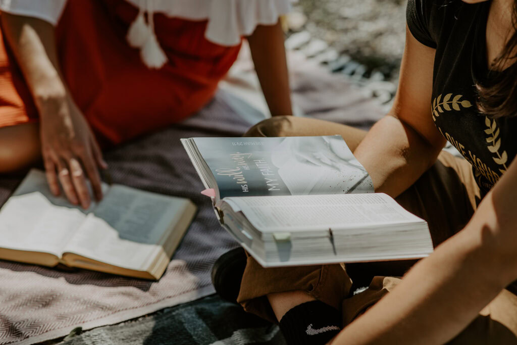 Zwei Frauen lesen in der Sonne auf einer Decke, eine hält ein offenes Buch.