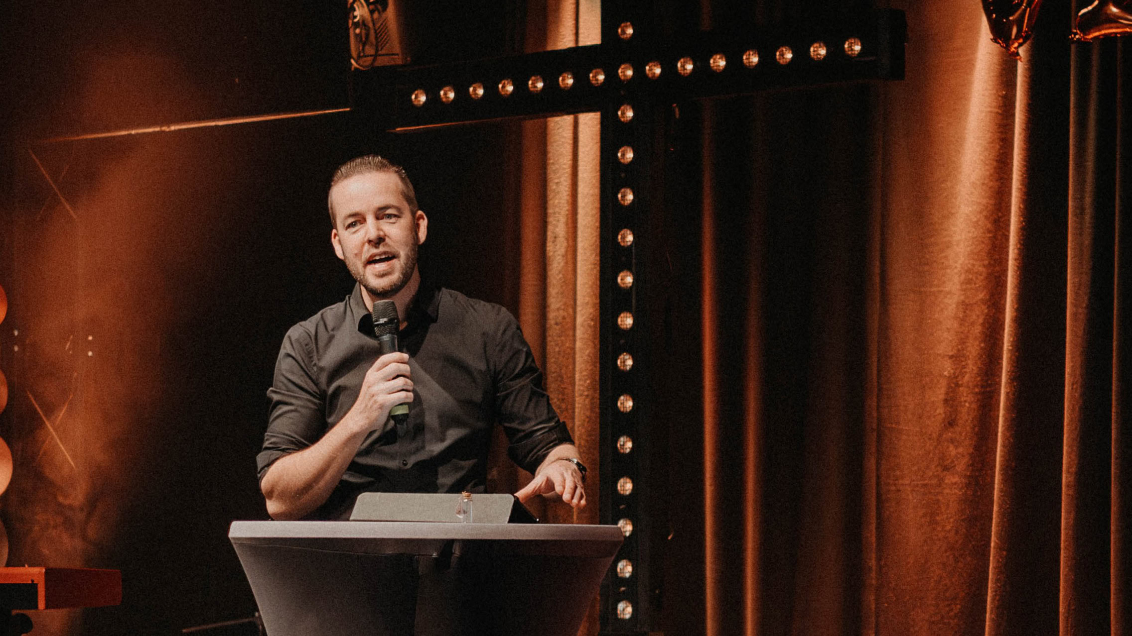 Ein Mann steht an einem Rednerpult auf einer Bühne, hält ein Mikrofon in der Hand und spricht.