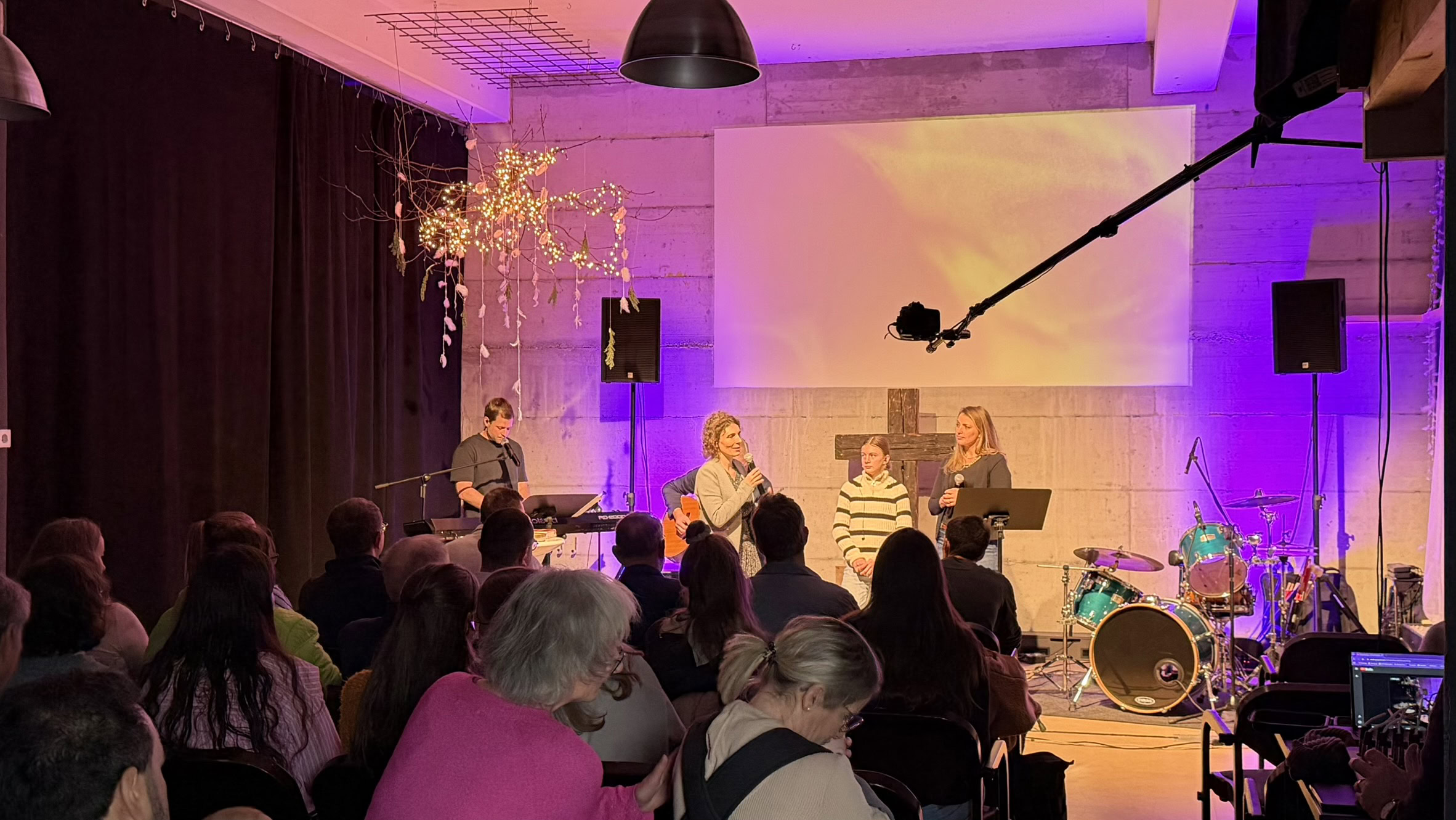 Drei Musiker auf einer Bühne mit Instrumenten, im Hintergrund eine große weiße Leinwand.