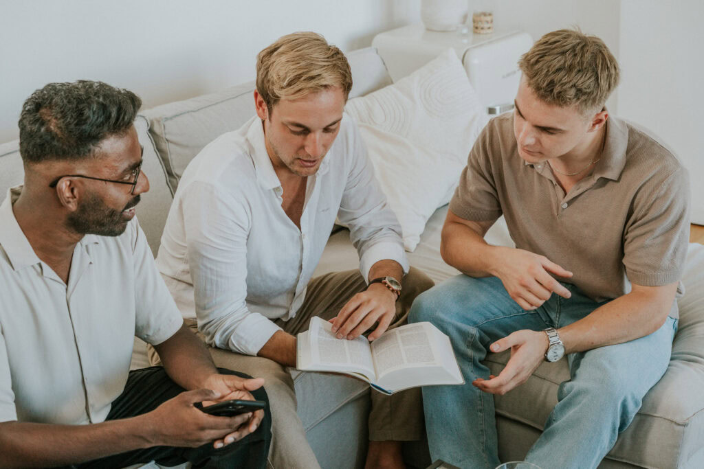 Drei Männer sitzen auf einem Sofa, diskutieren und schauen in ein Buch.