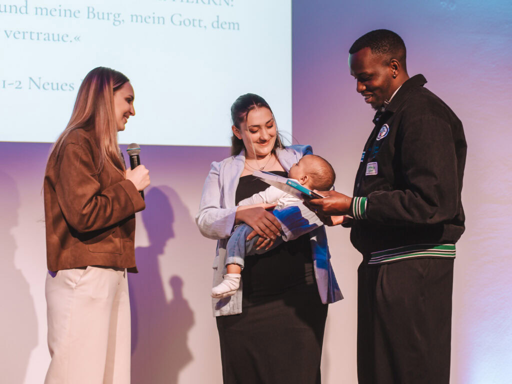 Frau hält Baby, während ein Mann ein Buch zeigt, neben einer Frau mit Mikrofon.