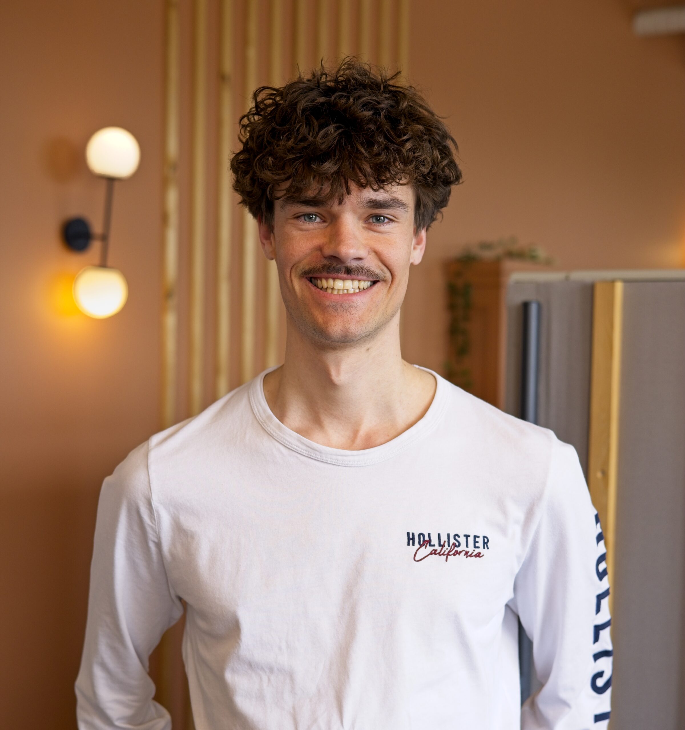 Ein junger Mann mit lockigem braunem Haar und Bart steht in einem Büro, trägt ein weißes T-Shirt mit.