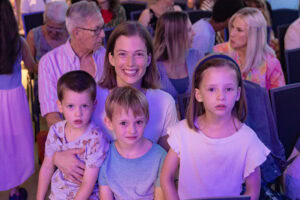 Frau mit drei Kindern sitzt in einem Saal mit anderen Menschen im Hintergrund.