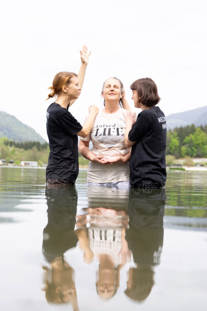 Drei Frauen stehen im Wasser, beten und heben die Hände, im Hintergrund Berge und Bäume.
