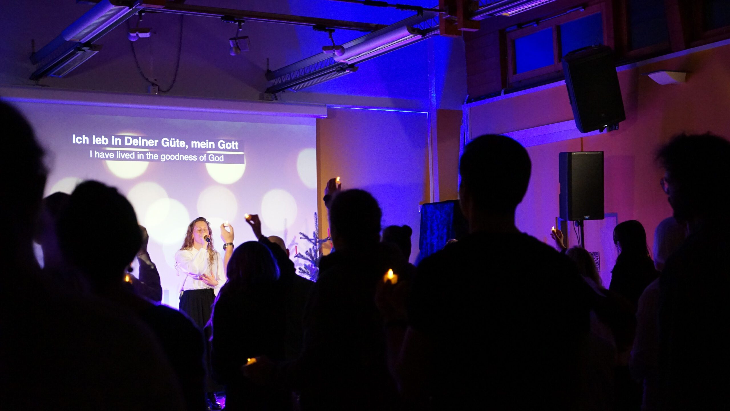 Menschen in dunklem Raum bei Gottesdienst, einige mit erhobener Hand.