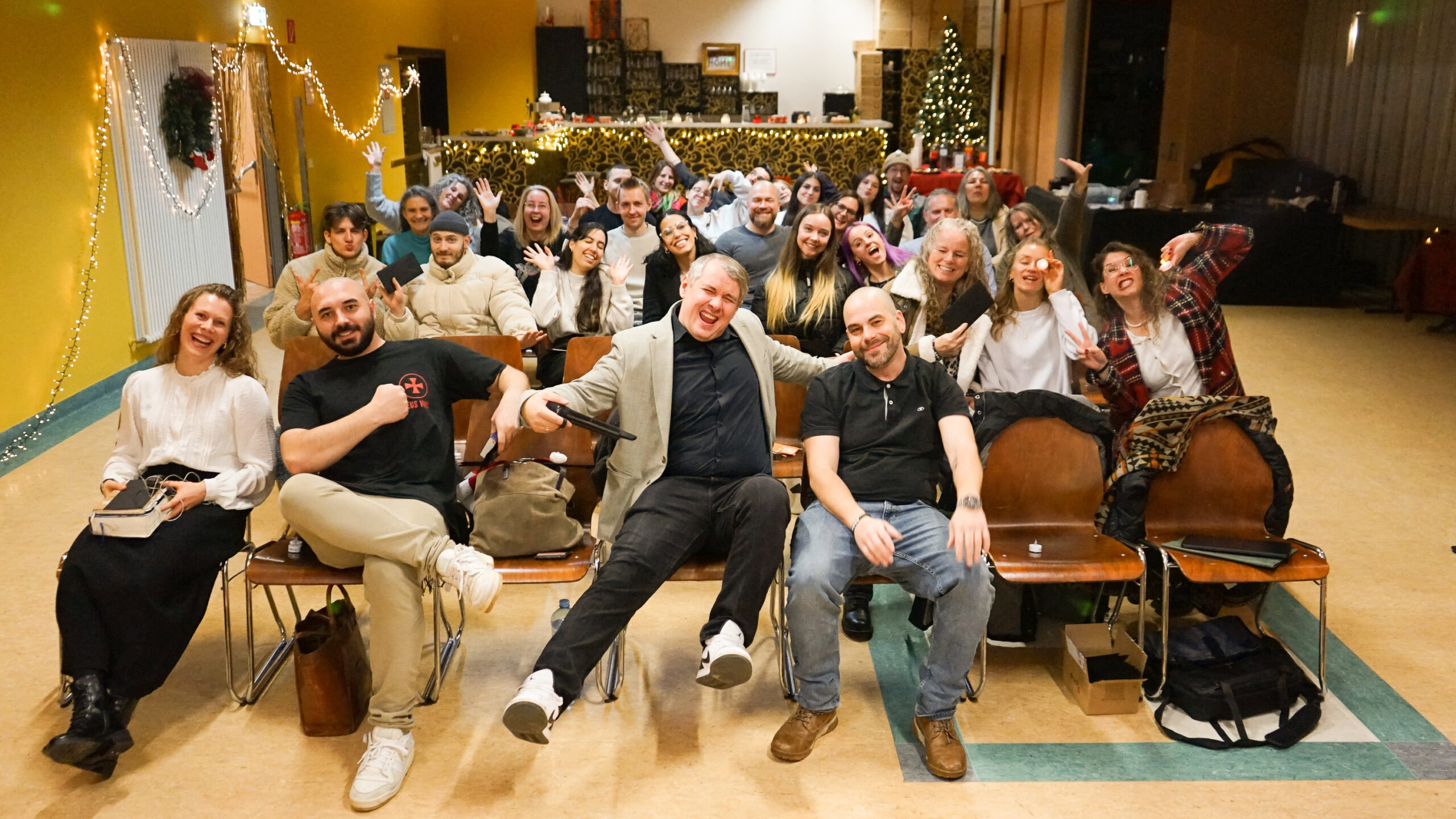 Gruppe von Menschen sitzt auf Bänken in einem festlich dekorierten Raum, einige lachen und winken.