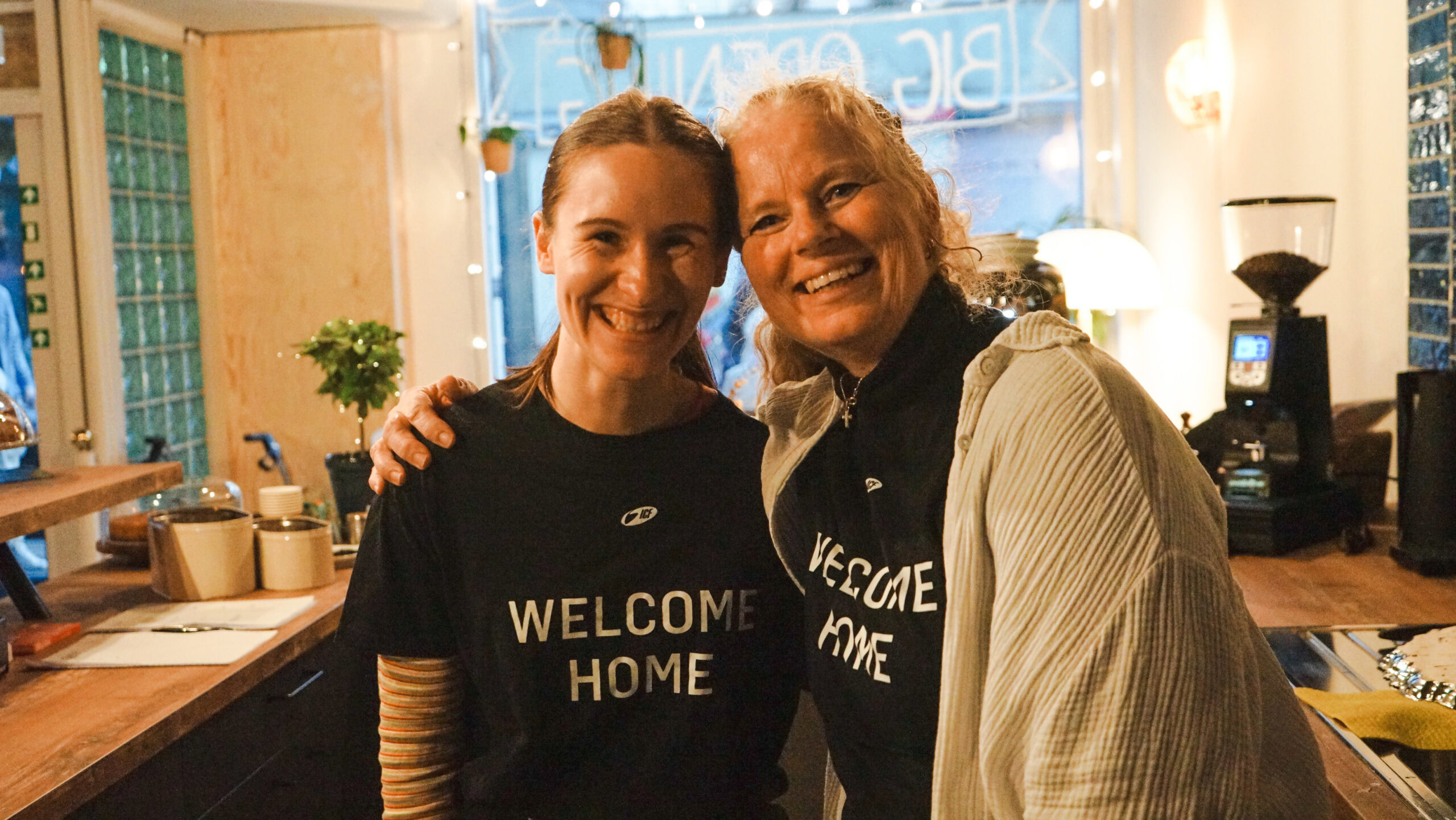 Zwei Frauen umarmen sich in einem Café, beide tragen T-Shirts mit "Welcome Home".