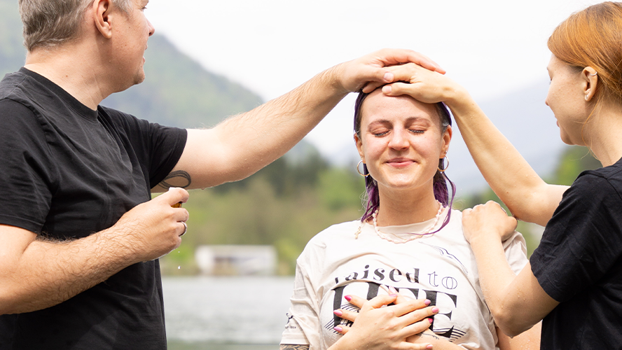 Zwei Erwachsene segnen eine junge Frau bei einer Taufe im Freien.