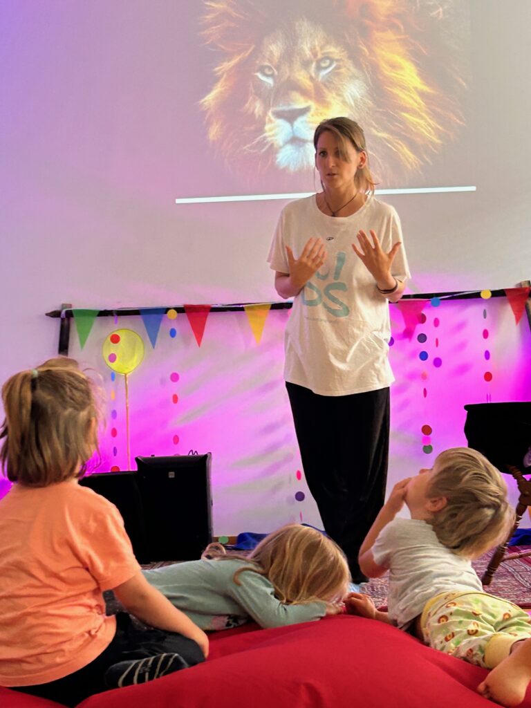 Frau steht und spricht vor Kindern im Raum, im Hintergrund ein Löwenbild.