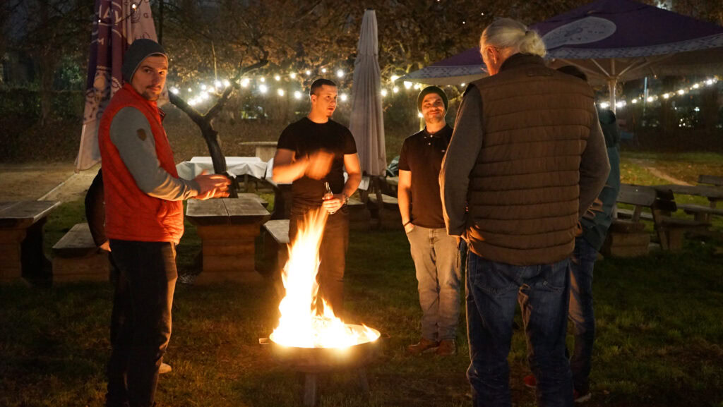 Fünf Menschen stehen im Kreis um eine Feuerstelle im Freien bei Nacht.