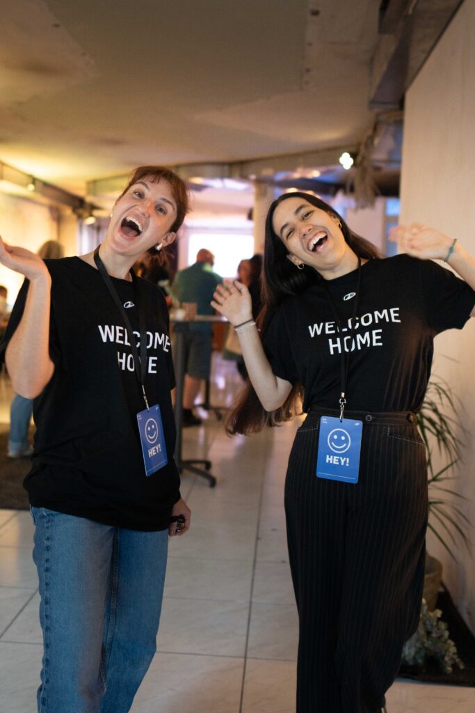 Zwei lachende Frauen tragen schwarze T-Shirts mit "Welcome Home" und haben blaue Hey-Aufkleber um de.