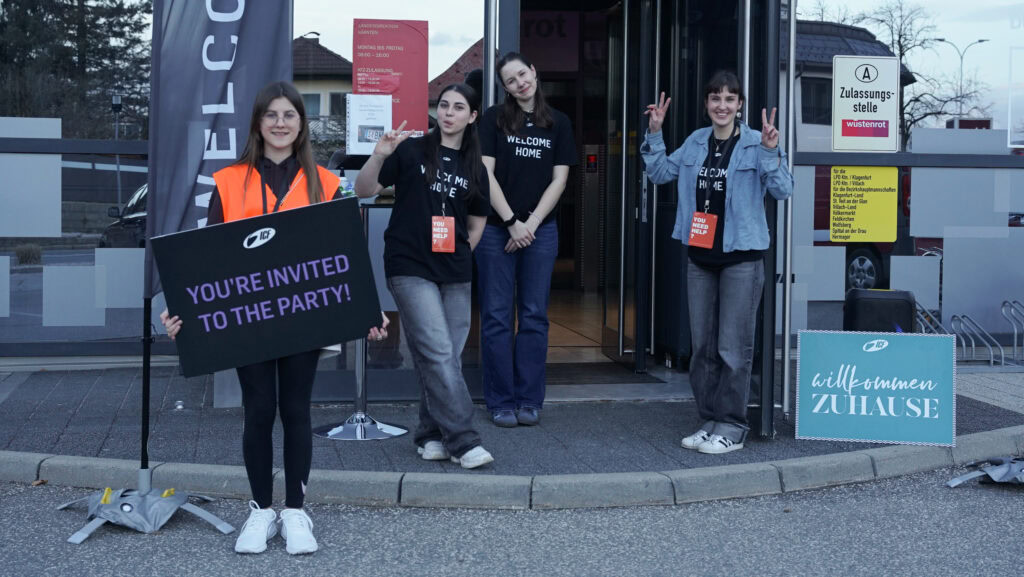 Vier junge Menschen stehen vor einem Gebäude, eines hält ein Schild mit "You're invited to the party.