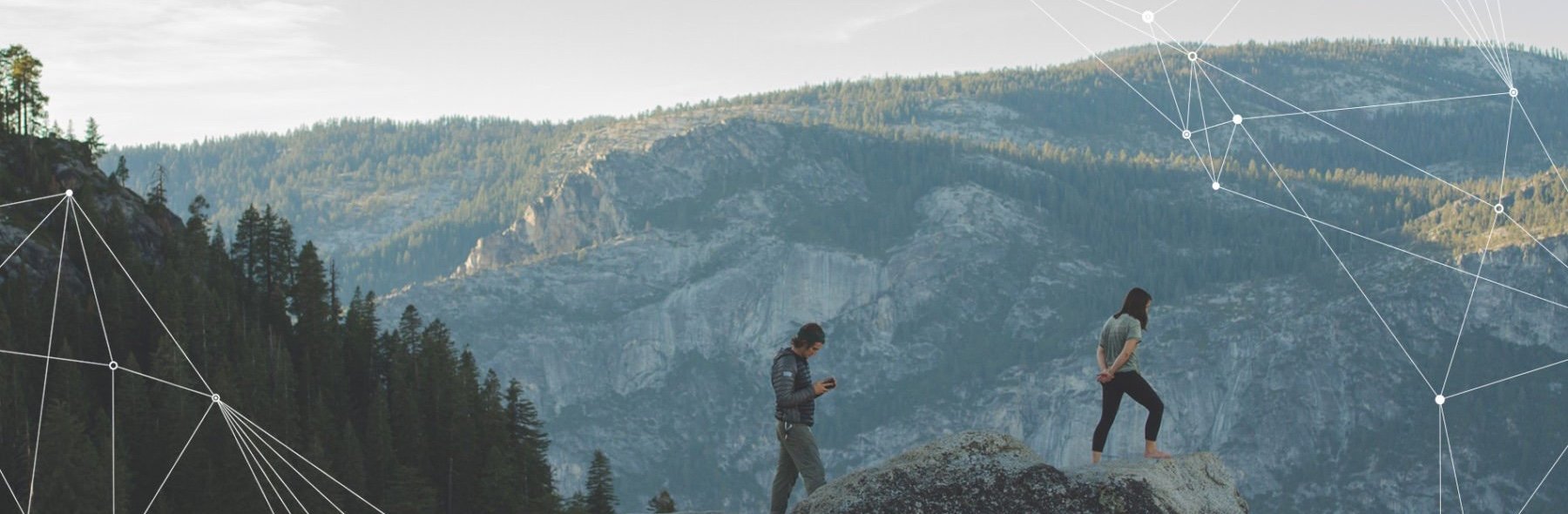 Zwei Personen stehen auf einem Felsen in einer Berglandschaft, eine Person schaut auf ein Smartphone.