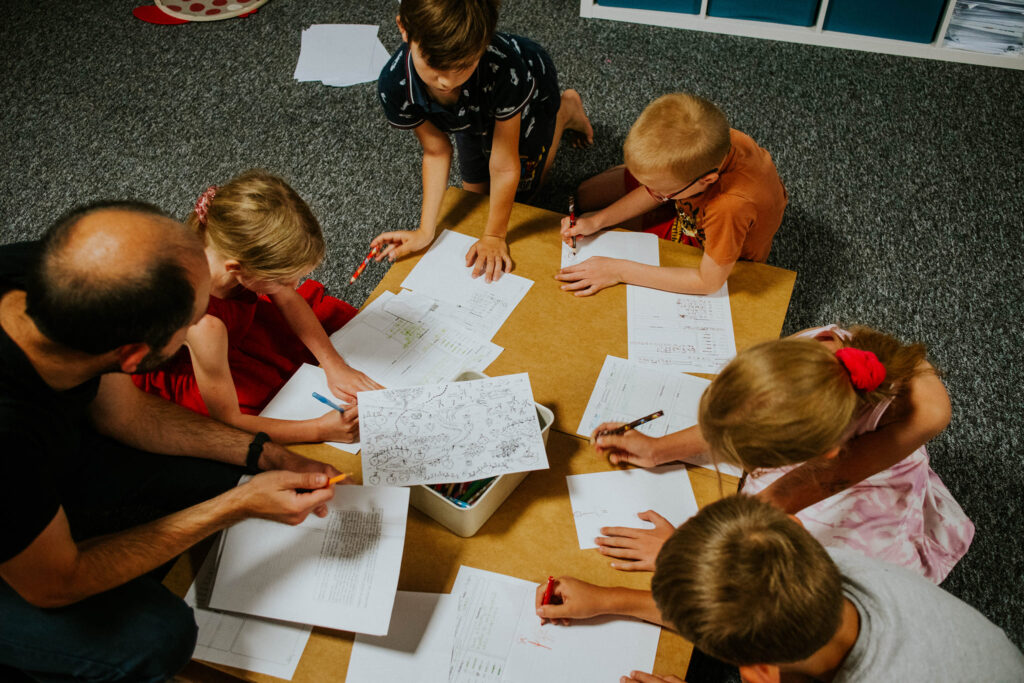 Six children and one adult gathered around a table with papers and notebooks.