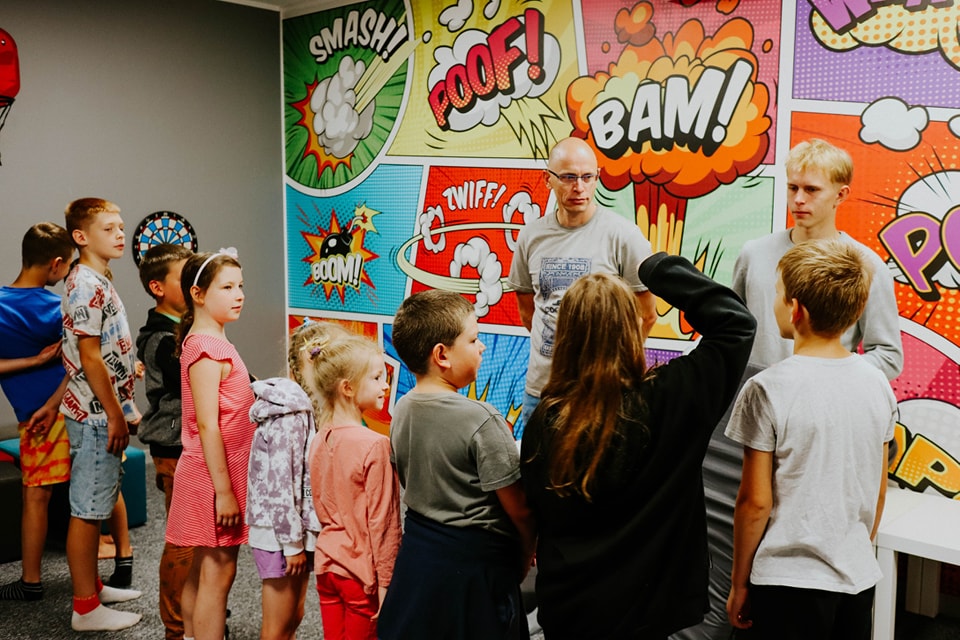 A group of children standing in front of a colorful comic-style wall with speech bubbles and explosi.