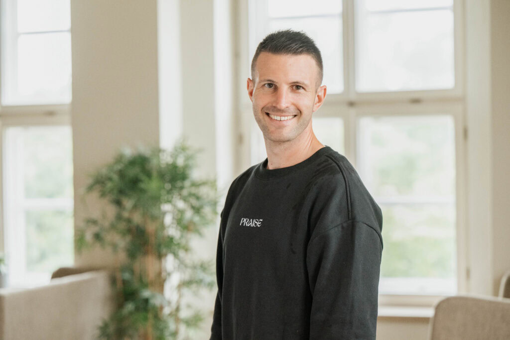 A man in a black sweater stands in a bright room with large windows.