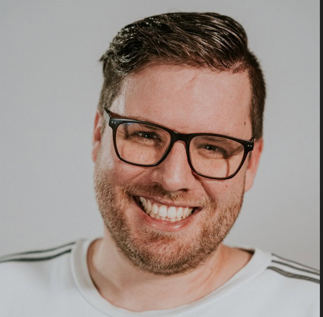 Mann mit Brille lächelt in die Kamera, kurze Haare, weißes T-Shirt, neutraler Hintergrund.