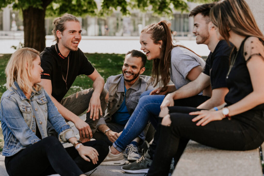 Several young people sit in a circle on the ground in the park and talk to each other.