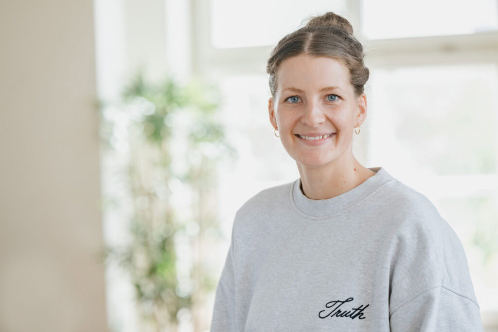 Bright young woman in a gray sweatshirt, smiling in front of a window in the background.