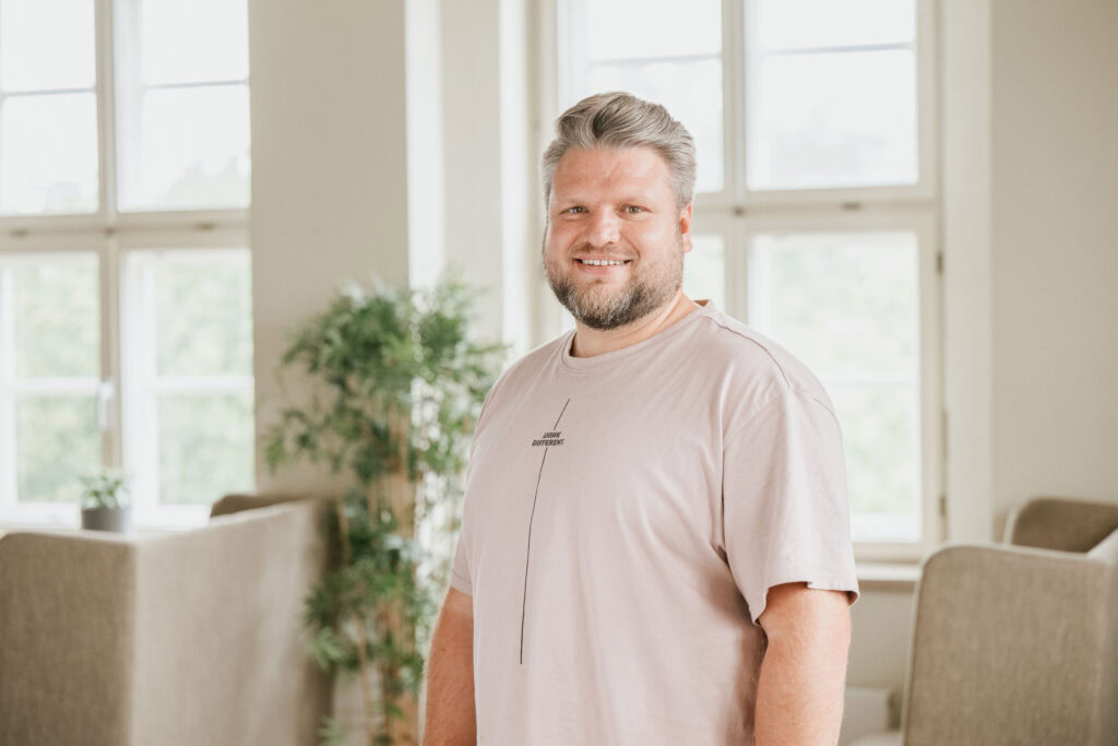 A smiling man with gray hair and a beard stands in a bright room.
