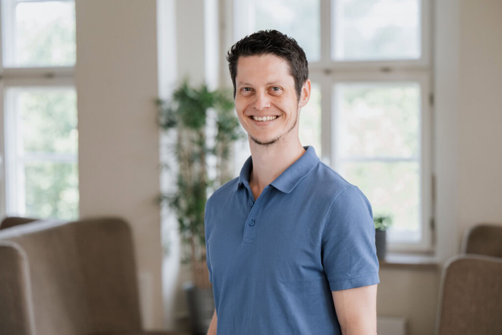 Man in a blue polo shirt stands in a bright living room with large windows.