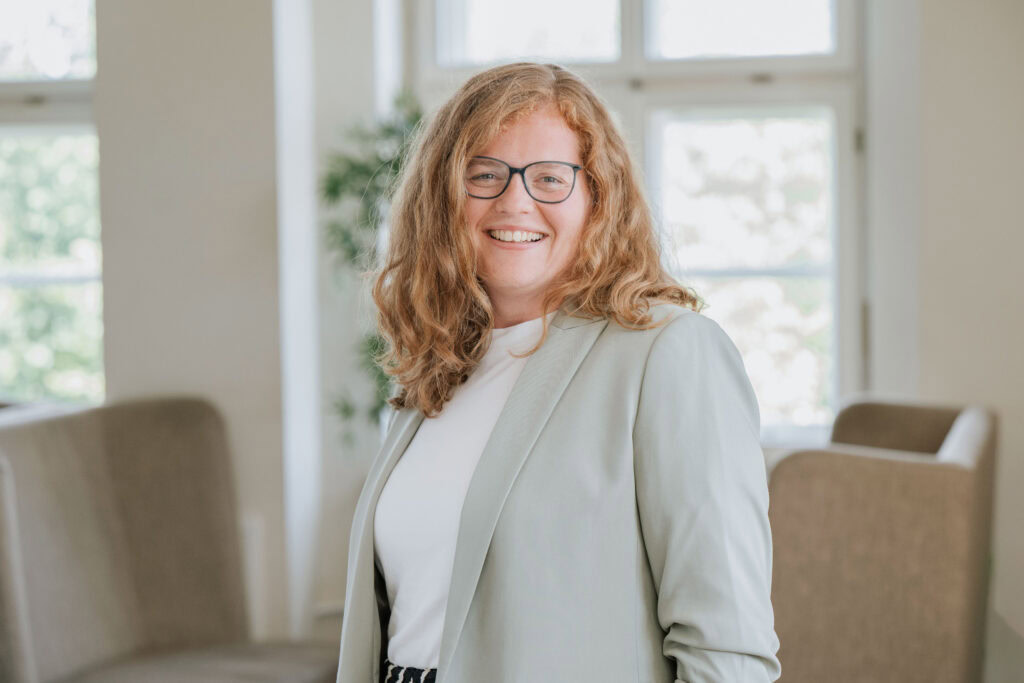 Woman with glasses and curly red hair smiles in a bright room.