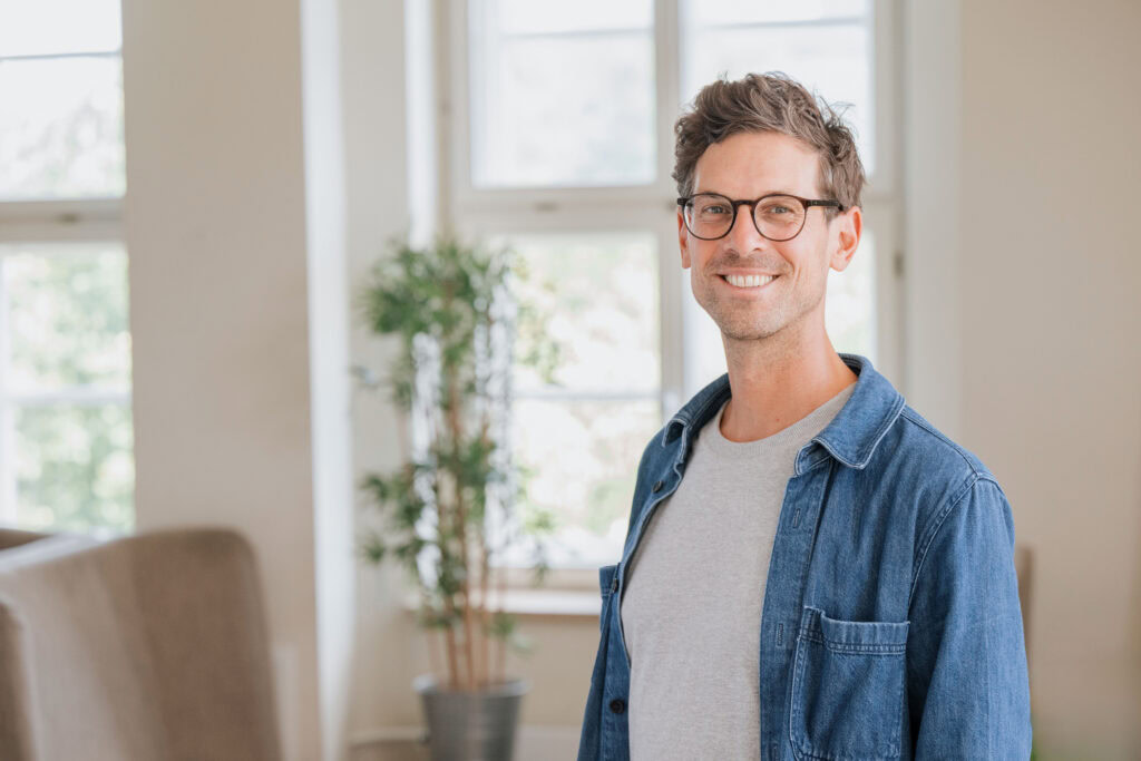 Man wearing glasses, a grey t-shirt and jeans in a bright room.