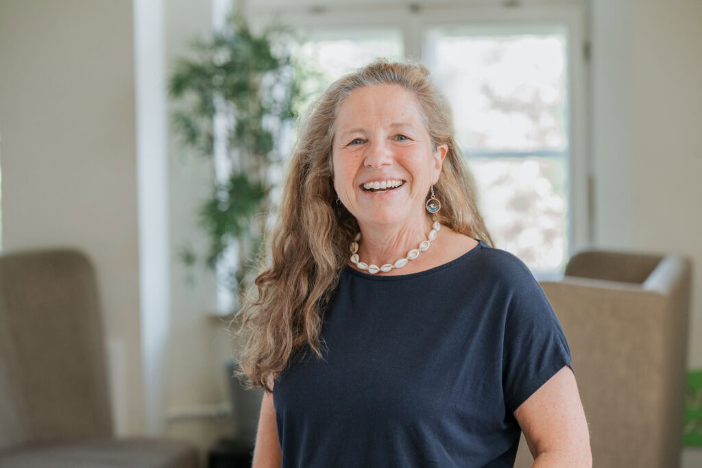 An older woman with long, curly hair, smiling in a blue T-shirt in front of a window.