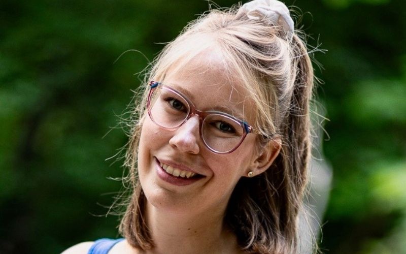 Young woman with glasses smiling in the park, green trees in the background.
