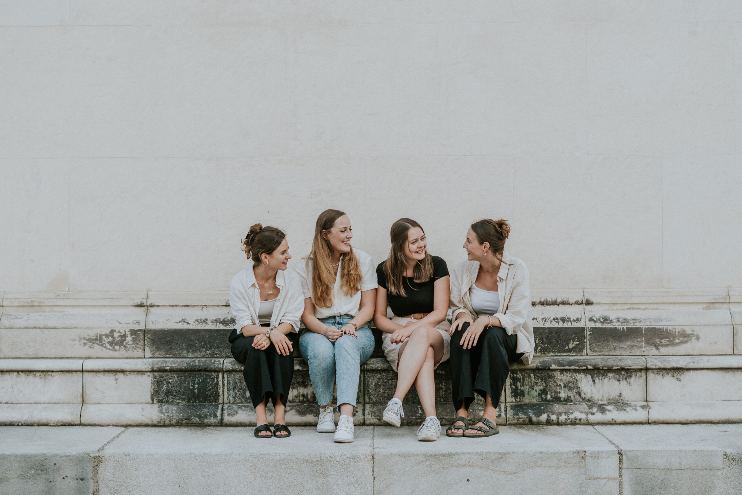 Vier junge Frauen sitzen auf einer Steinbank gegen eine weiße Wand und sprechen miteinander.
