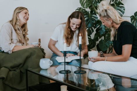 Drei Frauen sitzen auf einer Couch und einem Sessel, trinken Kaffee und lesen.
