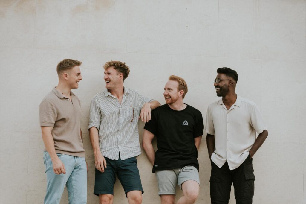 Four men stand in front of a white wall, laughing and talking.