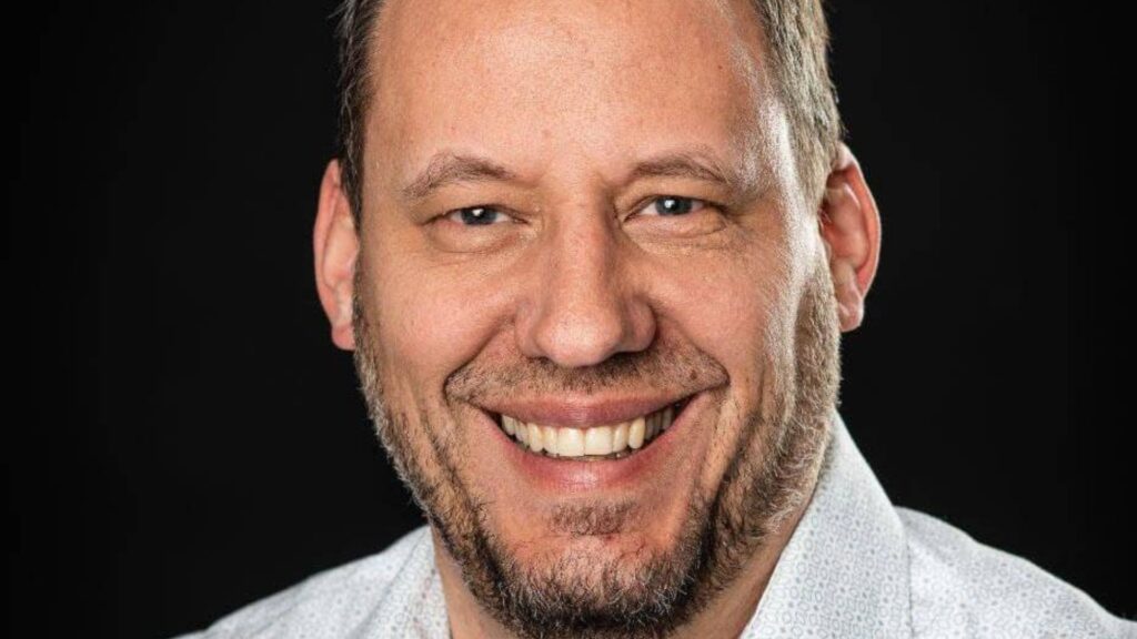Mann mit Bart und kurzen Haaren, lächelnd, in weißem Hemd.