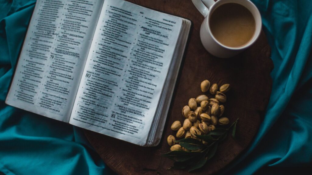 Ein offenes Buch mit Bibeltext liegt auf einer runden Holzplatte, daneben eine Tasse Kaffee und eine.