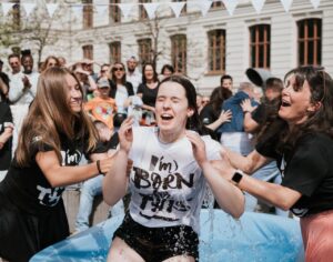 Several people are laughing and celebrating outdoors, one person gets wet in a pool of water.
