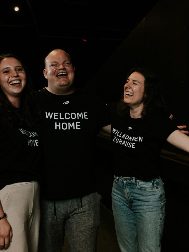 Drei Menschen lachen und umarmen sich, tragen schwarze T-Shirts mit "Welcome Home" und "Willkommen Z.