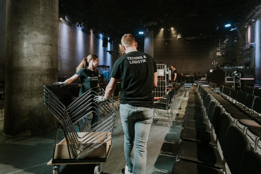 Two people carry chairs into an event room equipped with rows of chairs.