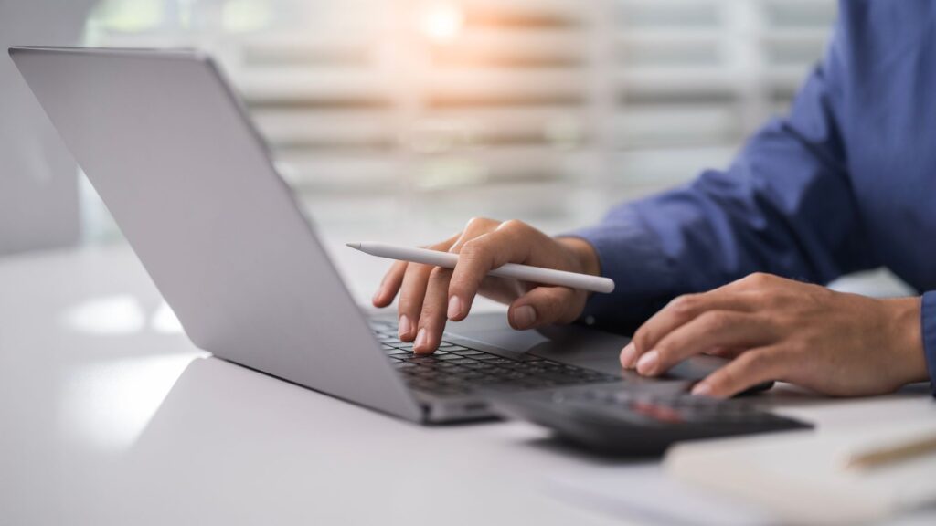 Person typing on laptop with pen in hand.