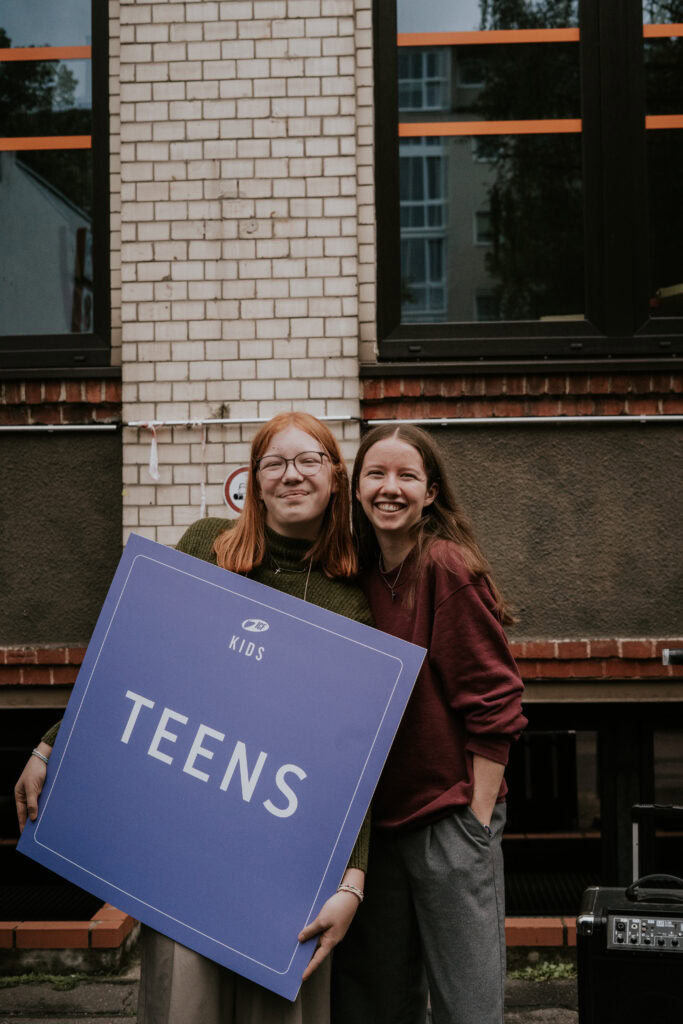 Zwei lachende Jugendliche halten ein großes Schild mit "TEENS" vor einem urbanen Hintergrund in Berlin, Deutschland.