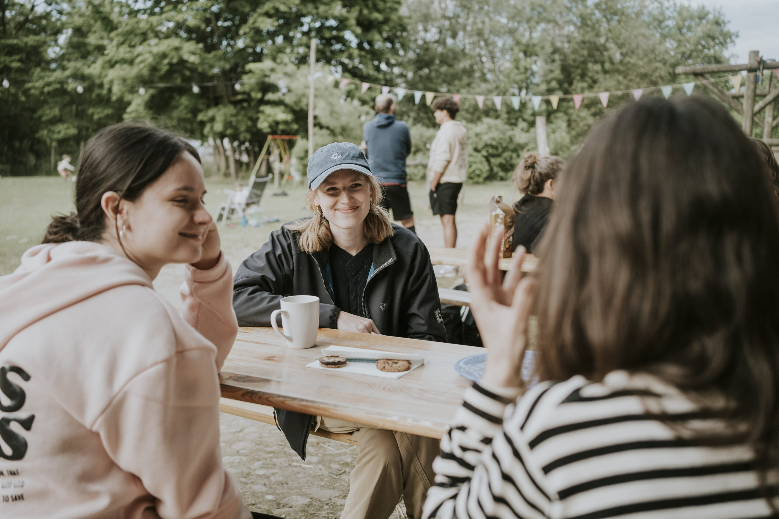 Drei Frauen sitzen an einem Holztisch im Freien, unterhalten sich.
