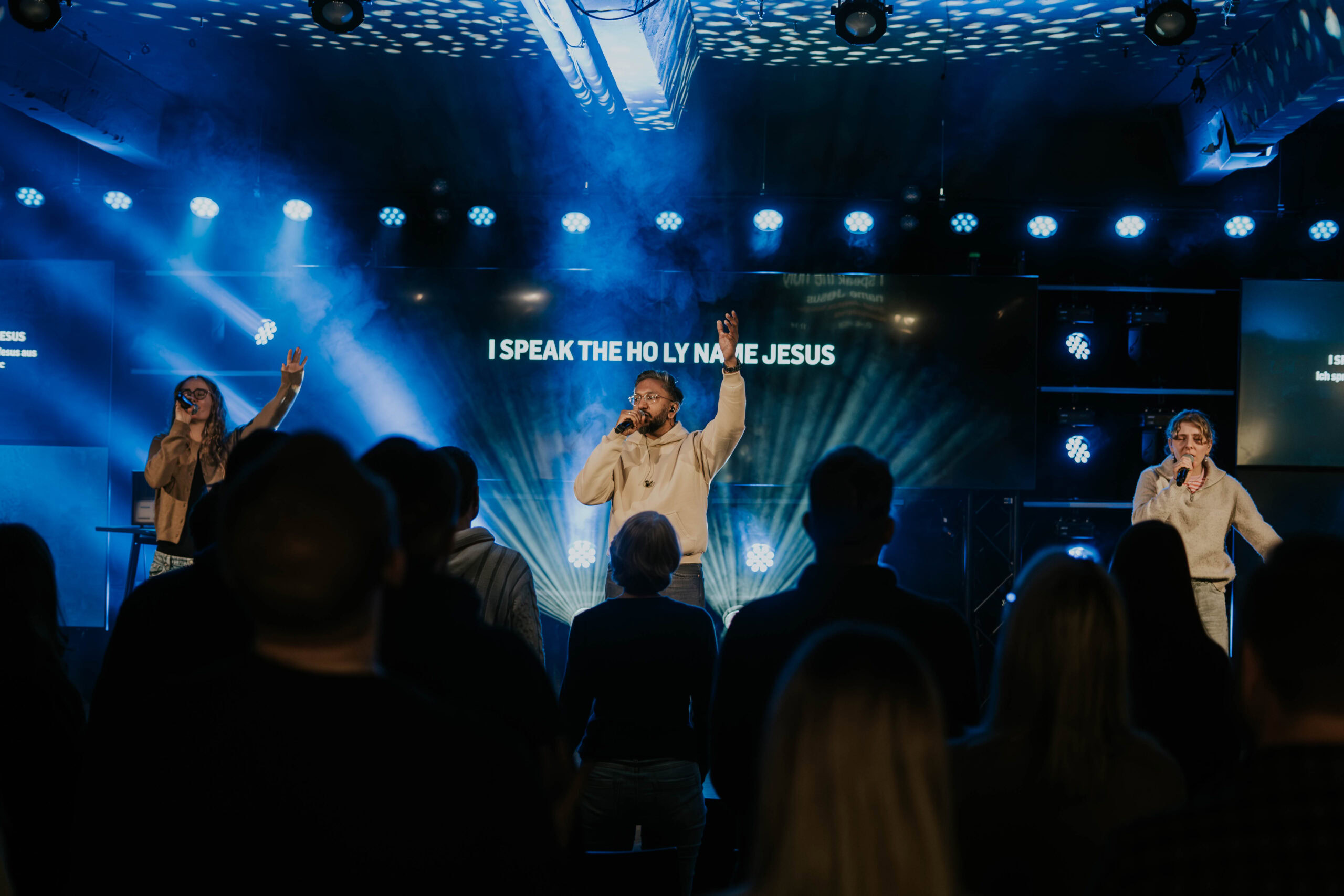 Menschen auf der Bühne beim Gottesdienst, Sänger mit Mikrofon, im Hintergrund Lichter und Projektion.