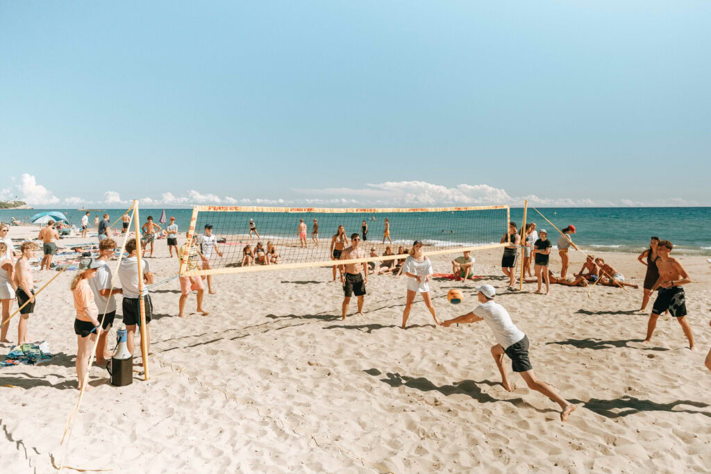 Kinder und Erwachsene beim Volleyball auf dem Sandstrand, blaues Meer im Hintergrund, sonniger Himmel, Ferienspaß, aktive Freizeit am Wasser.