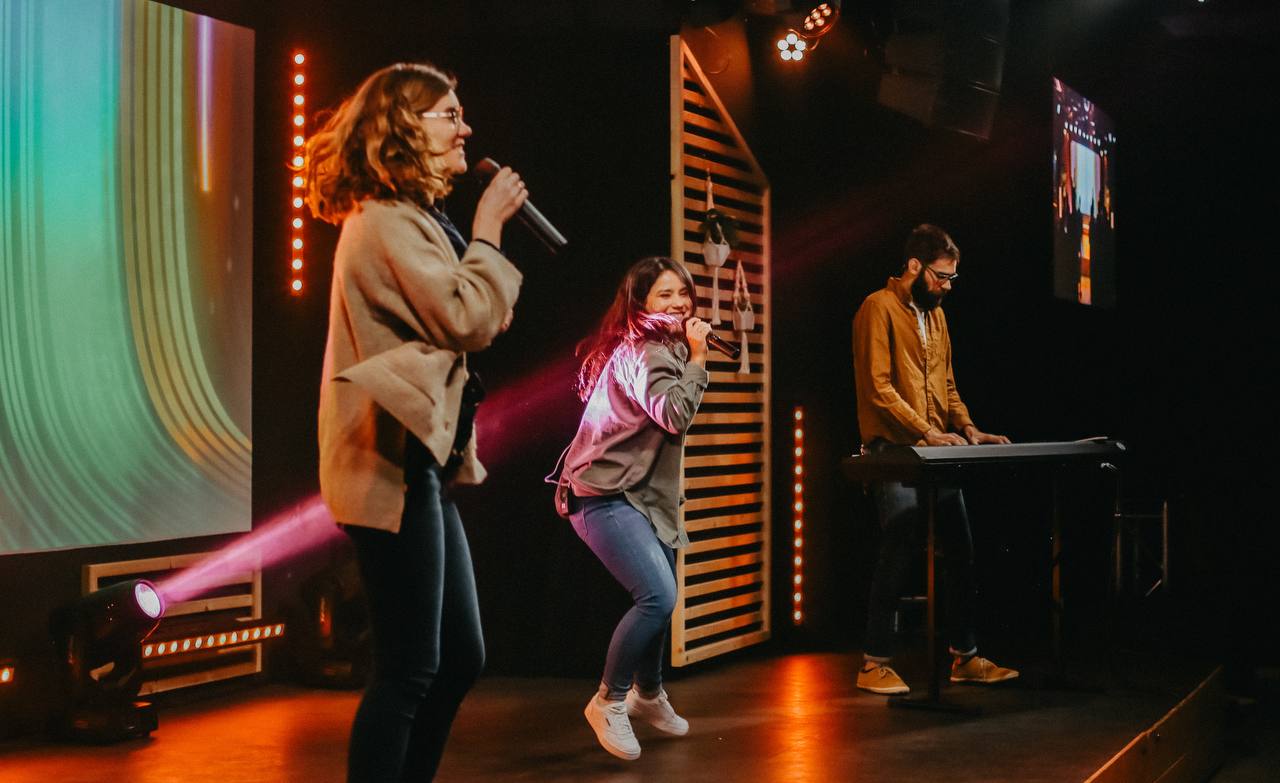 Worshipband steht und tanzt auf der Bühne und singt. Im Hintergrund ist eine Leinwand mit Liedtext.