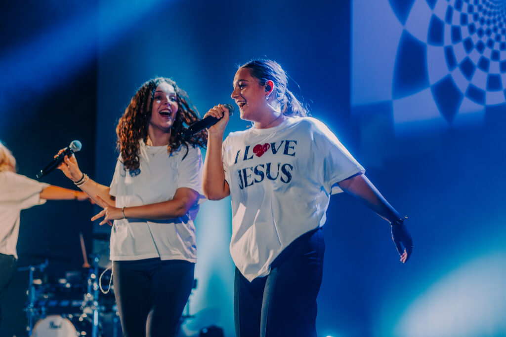 Zwei junge Frauen beim ICF Hamburg Jugendgottesdienst, lachen und singen auf der Bühne, umgeben von buntem Licht und moderner Bühnenbeleuchtung.