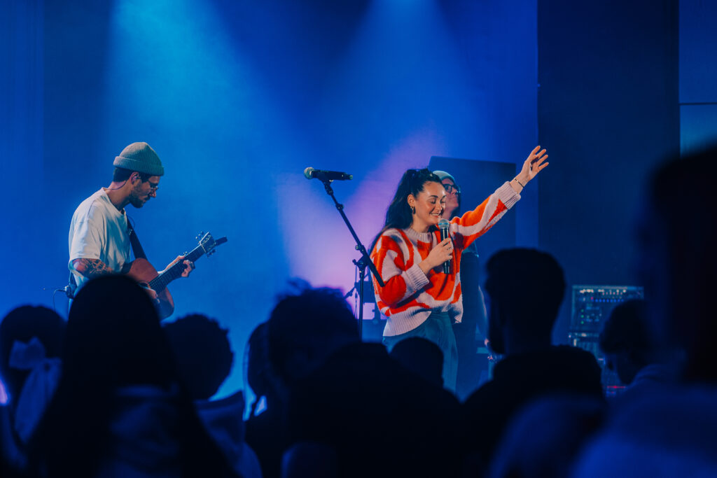 Junge Frau singt und preist Gott bei einem modernen Gottesdienst in Hamburg, begleitet von Gitarrenspielern, mit lebendiger Atmosphäre und engagierter Gemeinde.