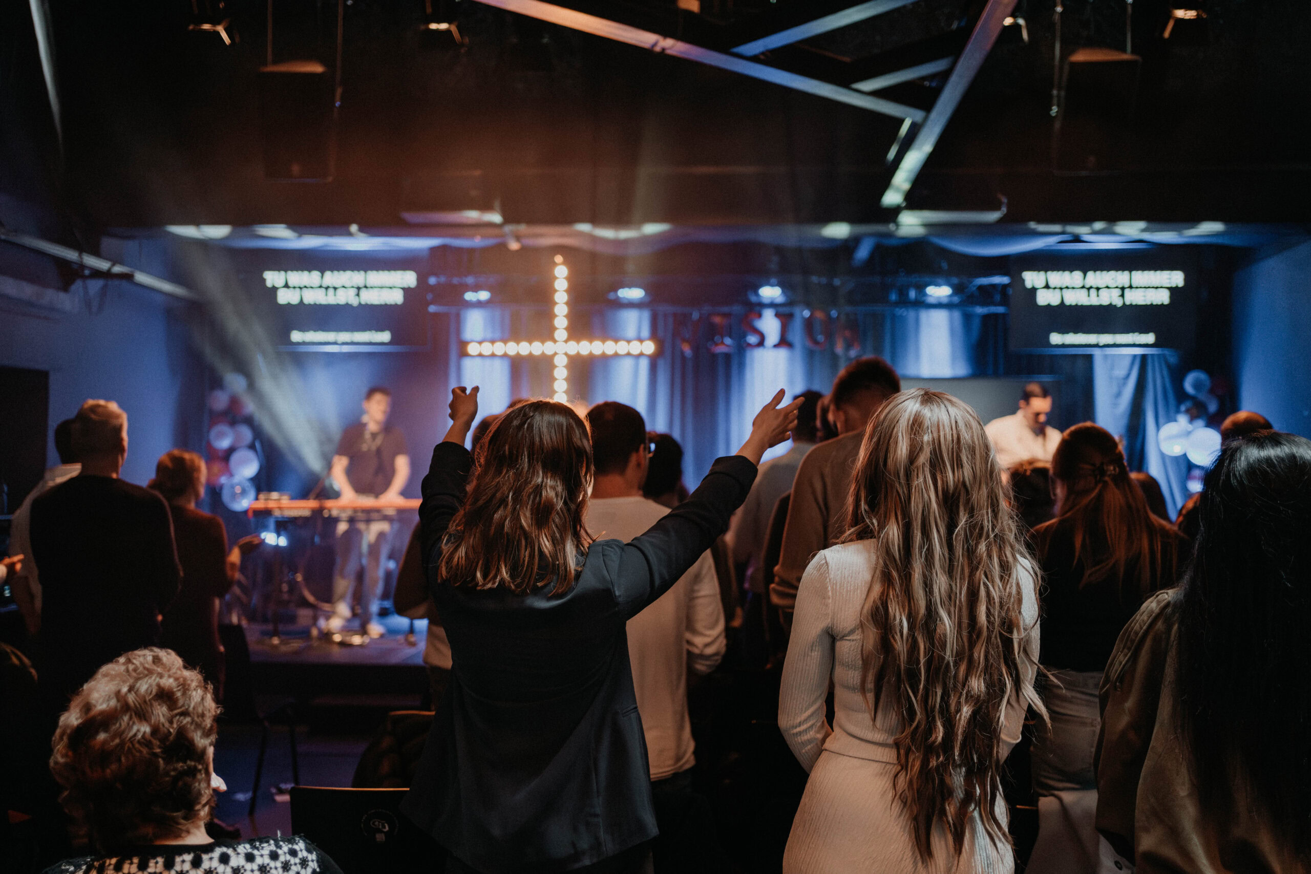 Menschen in einer Kirche während einer Feier, einige mit erhobenen Händen, im Hintergrund ein Kreuz.